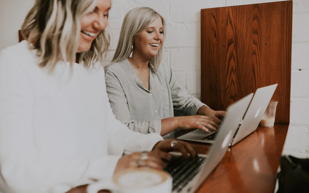Two women laughing and working side by side on laptops at a wooden counter in a bright café