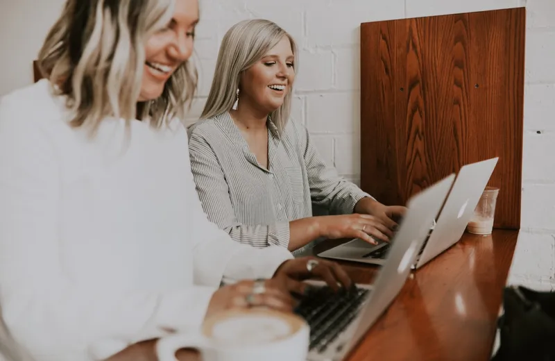 Two women laughing and working side by side on laptops at a wooden counter in a bright café