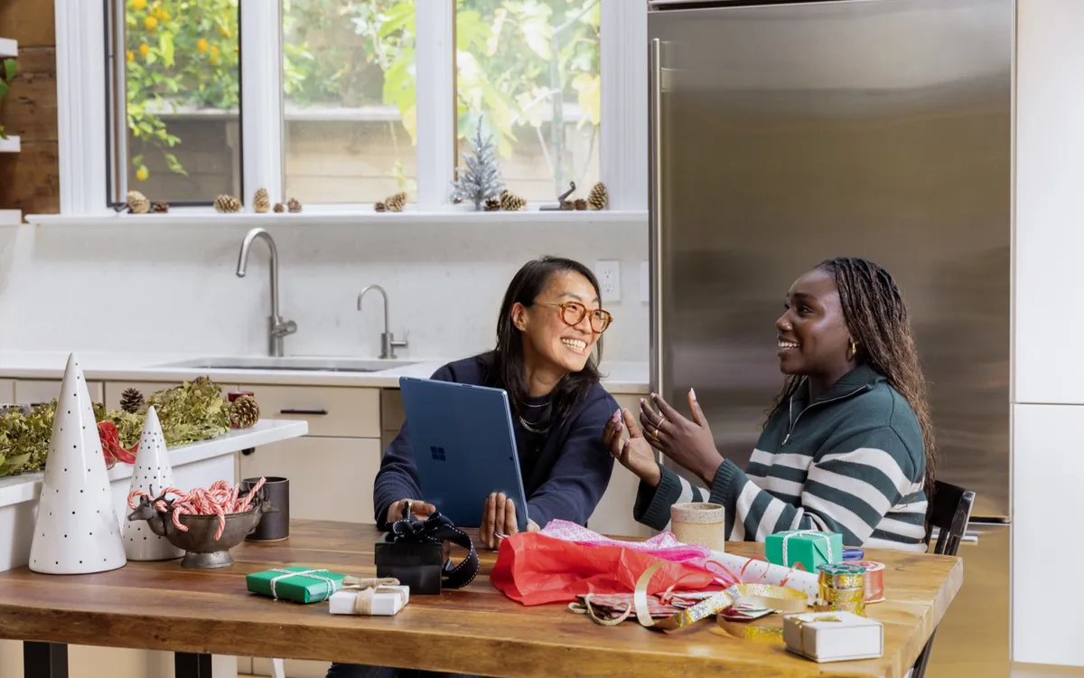 Two colleagues laughing together at a kitchen table over a tablet, with gifts and ribbon spread around them