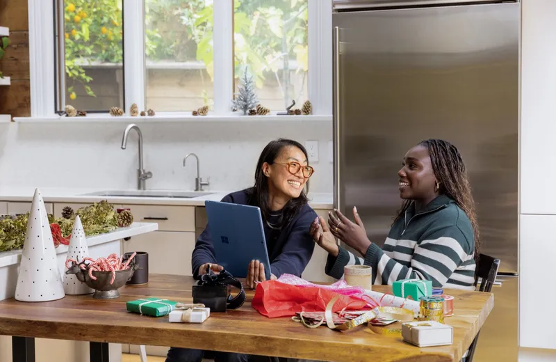 Two colleagues laughing together at a kitchen table over a tablet, with gifts and ribbon spread around them