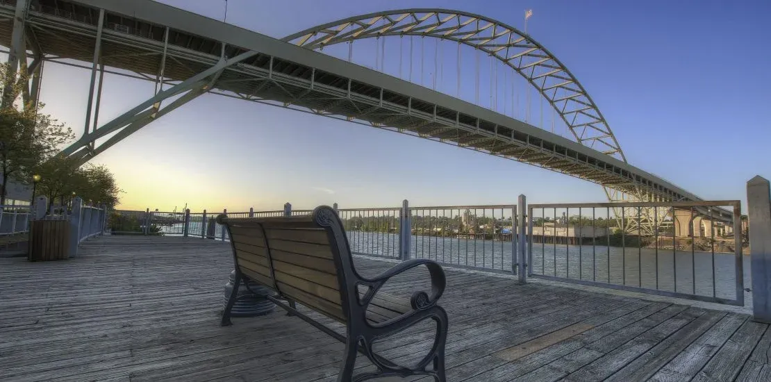 An empty wooden bench on a riverside boardwalk beneath Portland's Fremont Bridge at sunset