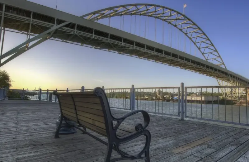 An empty wooden bench on a riverside boardwalk beneath Portland's Fremont Bridge at sunset