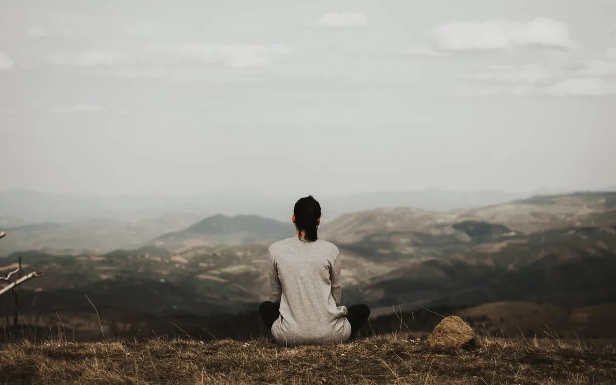 A person sitting cross-legged on a hilltop, facing away toward a hazy mountain range under an overcast sky