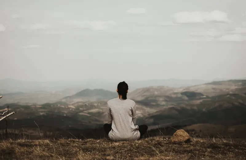 A person sitting cross-legged on a hilltop, facing away toward a hazy mountain range under an overcast sky