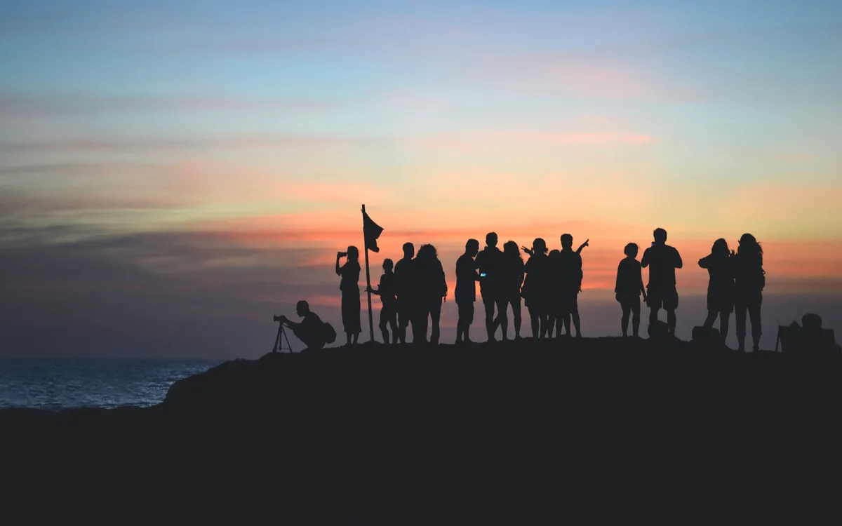 A group of people silhouetted against a sunset sky on a rocky shoreline, one holding a flag and one crouched behind a camera