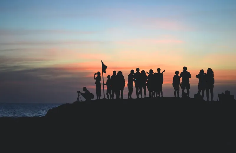 A group of people silhouetted against a sunset sky on a rocky shoreline, one holding a flag and one crouched behind a camera
