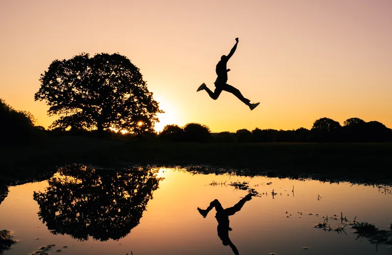 A person leaping over a puddle at sunset, silhouetted with one arm raised and reflected in the water beside a large tree
