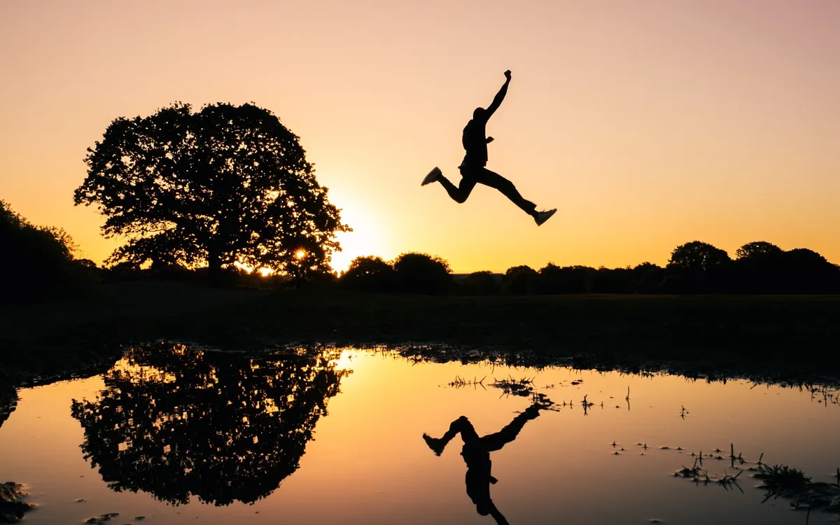 A person leaping over a puddle at sunset, silhouetted with one arm raised and reflected in the water beside a large tree